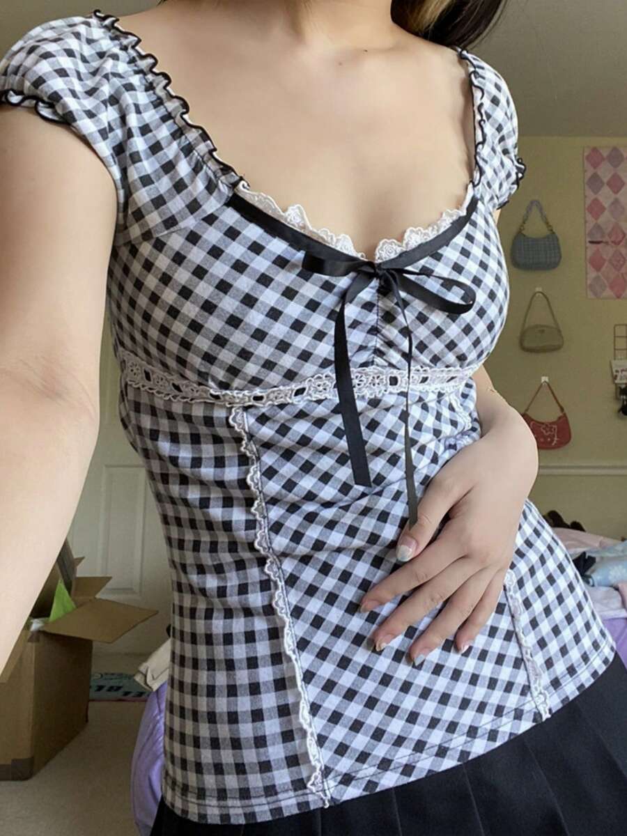 Women's black and white plaid short sleeve blouse featuring a tie-front front bow, contrast lace frill trim, raglan sleeves, and sweetheart neckline.