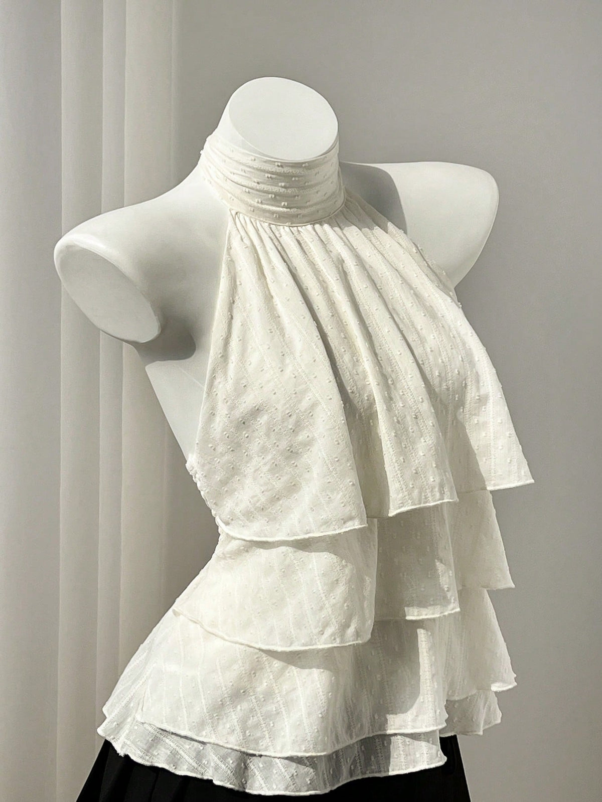 Women's white sleeveless halter neck top featuring pleated fabric, backless tie strap design, and a flared layered ruffle hem.