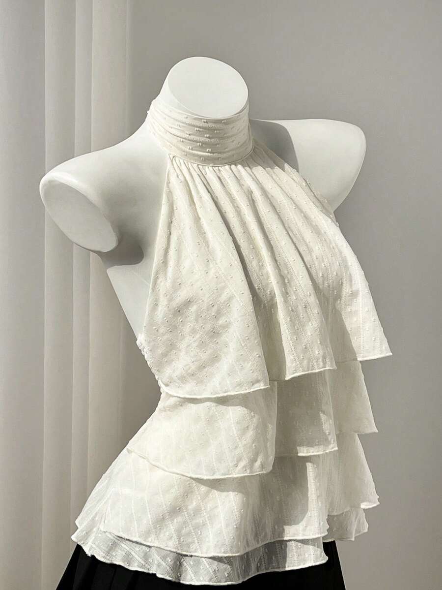 Women's white sleeveless halter neck top featuring pleated fabric, backless tie strap design, and a flared layered ruffle hem.