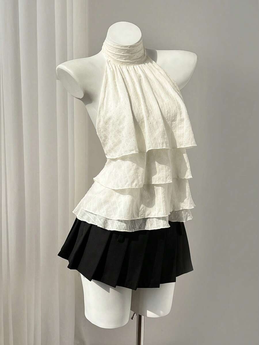 Women's white sleeveless halter neck top featuring pleated fabric, backless tie strap design, and a flared layered ruffle hem.
