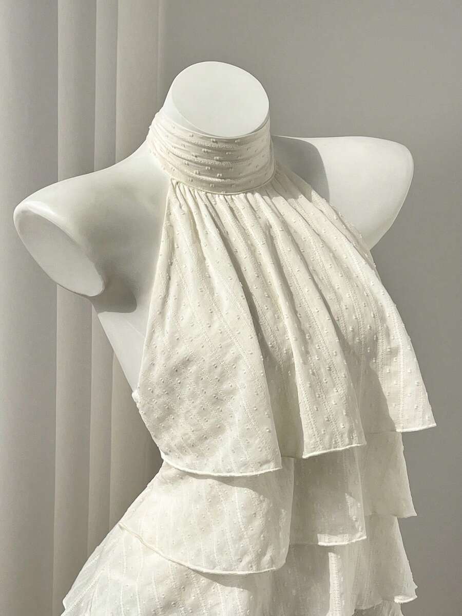 Women's white sleeveless halter neck top featuring pleated fabric, backless tie strap design, and a flared layered ruffle hem.