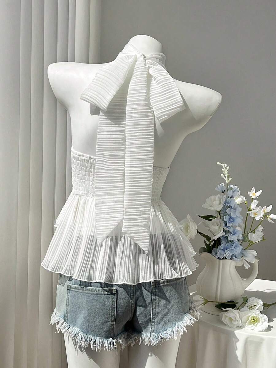 Women's white sleeveless halter neck top featuring pleated fabric, backless tie strap design, and a flared layered ruffle hem.