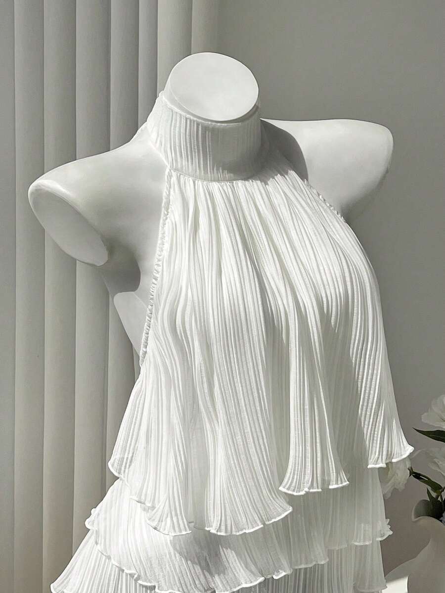 Women's white sleeveless halter neck top featuring pleated fabric, backless tie strap design, and a flared layered ruffle hem.