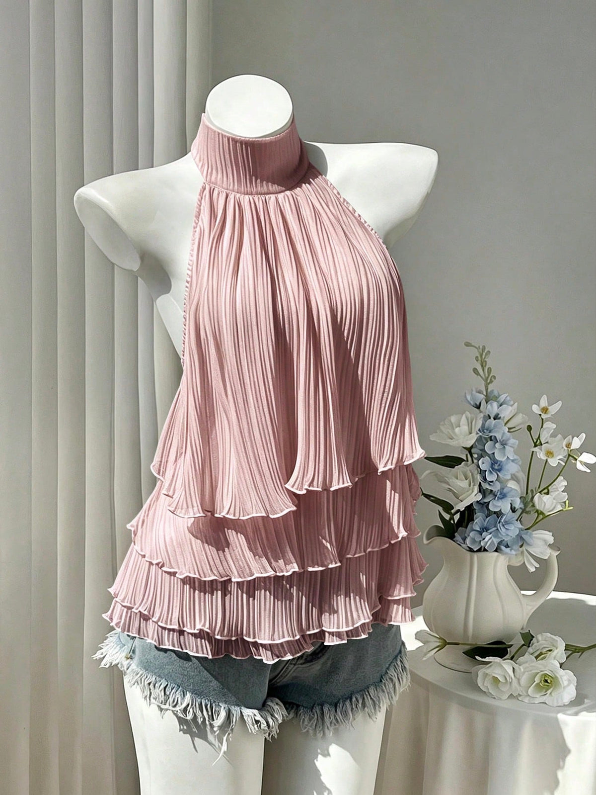 Women's white sleeveless halter neck top featuring pleated fabric, backless tie strap design, and a flared layered ruffle hem.