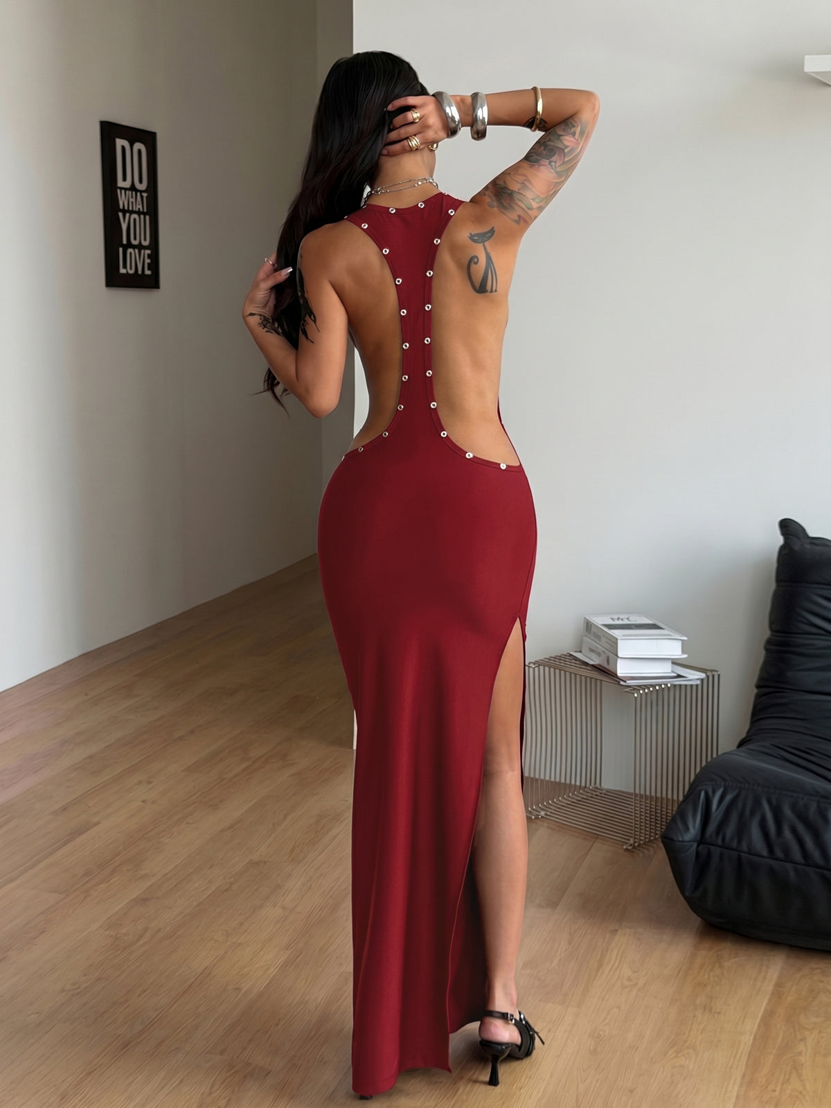 Front and side view of a women's black sleeveless bodycon maxi dress featuring a round neckline, open back, and high leg slit with metallic grommet eyelet details.